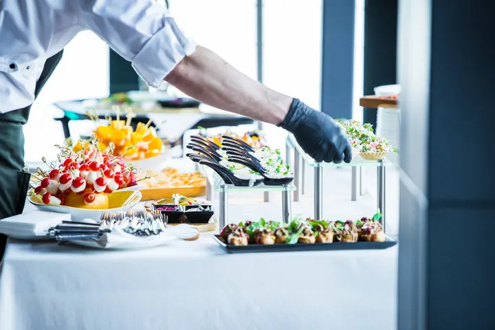 Close-up of a professional catering setup at a corporate event with gourmet appetizers, chef serving food with gloves, organized by Joyous Events